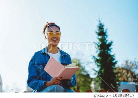 Positive female student in sunglasses enjoying studying on nature with textbook for education 108989417