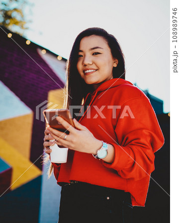 Positive young woman dressed in casual wear installing app on mobile phone while smiling at camera 108989746