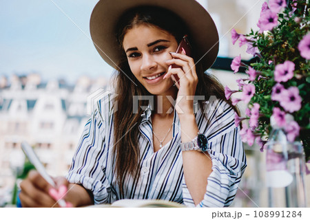 Attractive caucasian woman in hat looking at camera while talking on mobile phone sitting outdoors 108991284