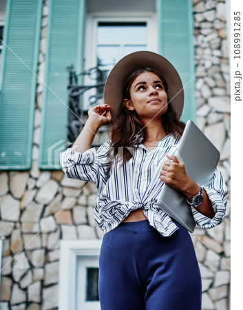 Beautiful young teen woman dressed in trendy apparel and hat standing on urban setting with book 108991285