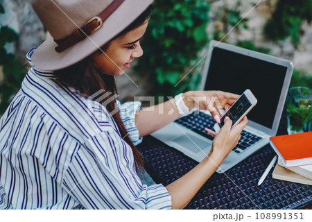 Positive hipster girl in hat checking new feed in social networks using mobile phone and 4G connection 108991351