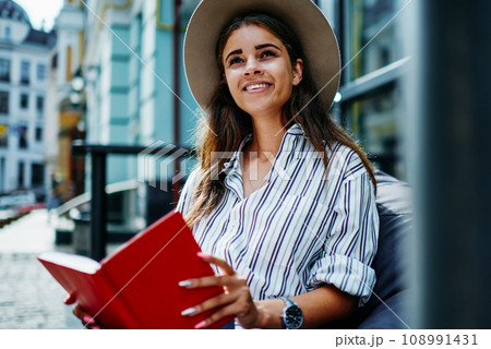 cheerful hipster girl in stylish hat spending free time on hobby holding interesting best seller 108991431