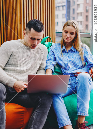 Young man and woman dressed in casual wear sitting on comfortable poufs watching film online Young man and woman dressed in casual wear sitting on comfortable poufs watching film online 108991936