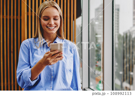 Positive young woman chatting online on cellular standing in office interior 108992113