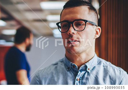 Serious young man in eyeglasses standing in office and communicating with colleague 108993035