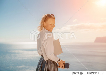 Freelance women sea working on a computer. Pretty middle aged woman with computer and phone outdoors with beautiful sea view. The concept of remote work. 108995192