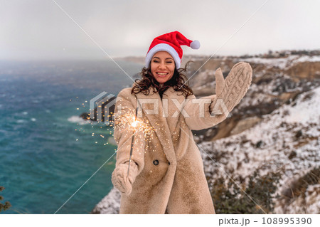 Outdoor winter portrait of elegant happy smiling woman in santa hat, light faux fur coat holding sparkler, posing against sea and snow background. 108995390