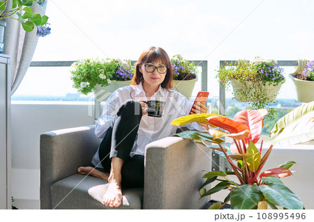 Middle aged happy woman sitting at home in chair with cup of smartphone Middle aged happy woman sitting at home in chair with cup of smartphone 108995496