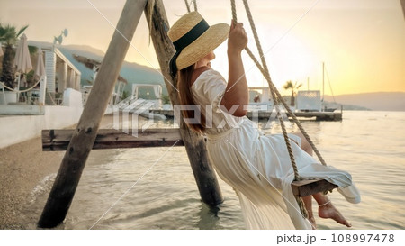 Sea beach where a woman in an elegant long dress gracefully sways on a rope swing at sunset. Tourism, the wonder of summertime, the exhilaration of freedom, and the journey of travel 108997478