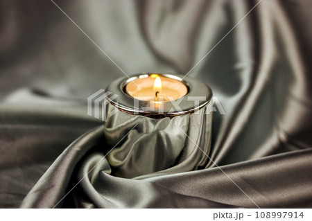 A burning candle in a metal candle holder on a table covered with a silver silk tablecloth. Luxurious stylish decor for modern interior A concept of tranquility, relaxation, meditation, self-knowledge 108997914