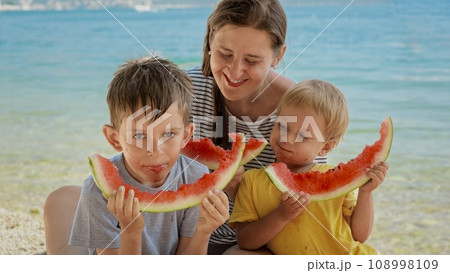 Joyful mother and her two sons enjoying eating sweet watermelon by the sea. Spirit of summertime, holiday celebrations, and the relaxation of parents enjoying quality time with their kids 108998109