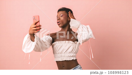 Selfie with phone of black transgender person in photo studio with pink background Selfie with phone of black transgender person in photo studio with pink background 108998123