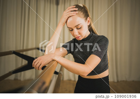 Upset ballerina in training suit stands near ballet barre and looks at the mirror in dance studio. Woman prepares for performance. Ballet dancer doing gymnastic exercises. Classical ballet school 109001936