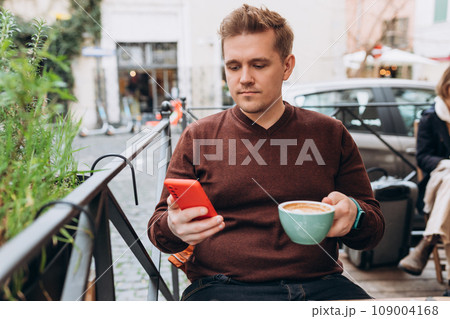 Unshaven happy man using mobile phone while drinking coffee at cafe. Smiling hipster guy checking email while messaging via cellphone technology 109004168