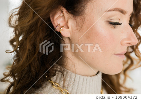 Cropped close-up shot of a young woman with two asymmetrical golden ear cuffs. Female with golden ear cuffs, side view. 109004372
