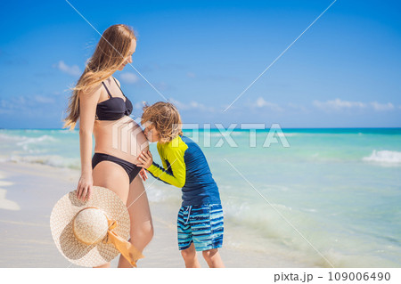 A radiant pregnant mother and her excited son share a tender moment on a serene, snow-white beach, celebrating family love amidst nature's beauty 109006490