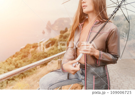 Woman rain umbrella. Happy woman portrait wearing a raincoat with transparent umbrella outdoors on rainy day in park near sea. Girl on the nature on rainy overcast day. 109006725