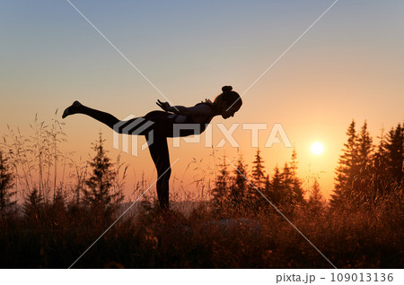 Young woman doing warrior 3 pose at fresh air. Silhouette of sporty female practicing yoga in mountains at sunrise. Exercising outdoor at early morning. Young woman doing warrior 3 pose at fresh air. Silhouette of sporty female practicing yoga in mountains at sunrise. Exercising outdoor at early morning. 109013136