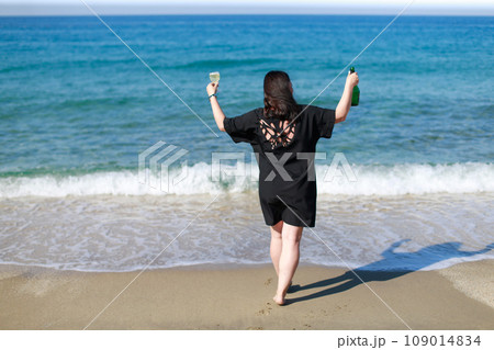 A girl in a black dress with champagne and a glass is standing on the seashore A girl in a black dress with champagne and a glass is standing on the seashore 109014834