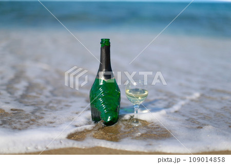 A bottle of champagne with a poured glass is standing on the seashore 109014858