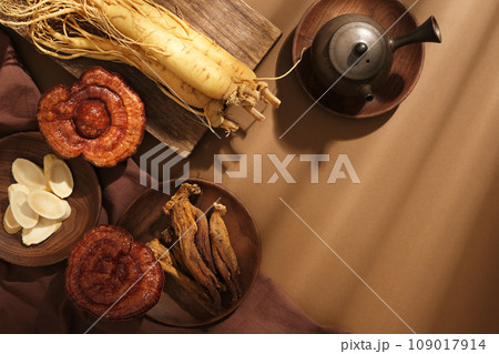 On a brown background, fresh ginseng roots, lingzhi mushrooms and red ginseng displayed on wooden tray and decorated with teapot. Blank space for the presentation product and design On a brown background, fresh ginseng roots, lingzhi mushrooms and red ginseng displayed on wooden tray and decorated with teapot. Blank space for the presentation product and design 109017914