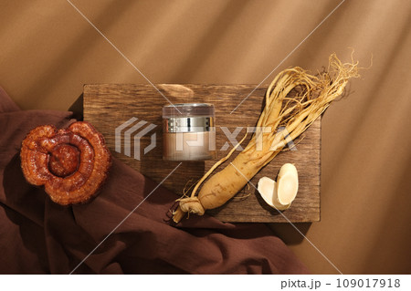 On a wooden podium, a cosmetic jar decorated with root and slices of ginseng, lingzhi mushroom and brown cloth on brown background. Concept for cosmetics extracted from rare herbs. Top view  109017918