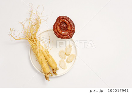 On a white background, fresh roots and slices of ginseng on white round ceramic dish decorated with lingzhi mushroom. Minimal concept for advertising product with rare herbal ingredient 109017941