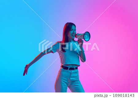 Young woman in casual clothes talking in megaphone against gradient pink blue studio background in neon light 109018143