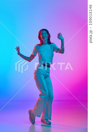 Young beautiful woman in casual clothes listening to music in headphones and dancing against gradient pink blue studio background in neon light 109018146