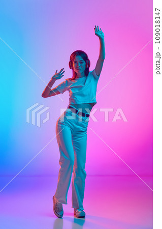 Young beautiful woman in casual clothes listening to music in headphones and dancing against gradient pink blue studio background in neon light Young beautiful woman in casual clothes listening to music in headphones and dancing against gradient pink blue studio background in neon light 109018147