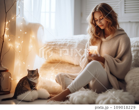 Young pretty woman dressed in cozy warm knitted sweater relaxing at her home with ginger cat, beautiful lady in white winter apartment interior decorated with candles and soft blankets 109018229
