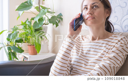 Brunette woman talking on the phone while sitting on the couch at home. 109018848