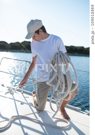 Young guy in white shirt and cap on the yacht fixing a rope Young guy in white shirt and cap on the yacht fixing a rope 109018869