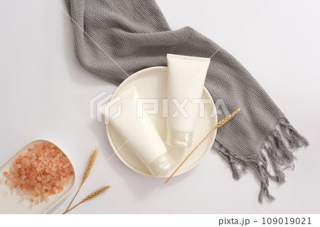 Round shaped dish featured two white tubes without label. A dish of pink himalayan salt displayed with a scarf. Daily self care routine concept Round shaped dish featured two white tubes without label. A dish of pink himalayan salt displayed with a scarf. Daily self care routine concept 109019021