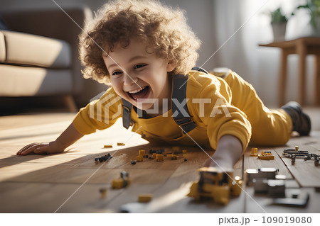 Happy child in yellow overalls with wavy hair plays with colorful toy blocks on the floor in a bright living room. Educational game for kids 109019080