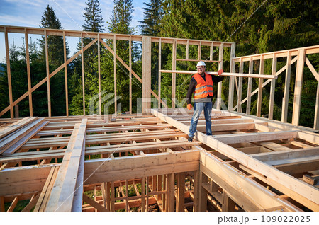 Carpenter builds wooden frame house near woods. Man holds large beam on shoulder, wearing work clothes and helmet. Concept of modern and sustainable building. 109022025