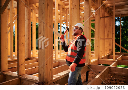 Carpenter constructing wooden frame house. Bearded man with glasses is hammering nails into the structure, wearing protective helmet and construction vest. Concept of modern ecological construction. Carpenter constructing wooden frame house. Bearded man with glasses is hammering nails into the structure, wearing protective helmet and construction vest. Concept of modern ecological construction. 109022028