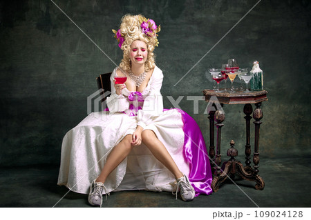 Upset, crying in despair queen, medieval person in old-fashioned dress and modern shoes sitting holding cocktail against vintage background. 109024128
