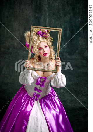 Woman dressed like classic historical character, in old-fashioned dress holding retro frame and grimacing looking at camera over vintage background. 109024144