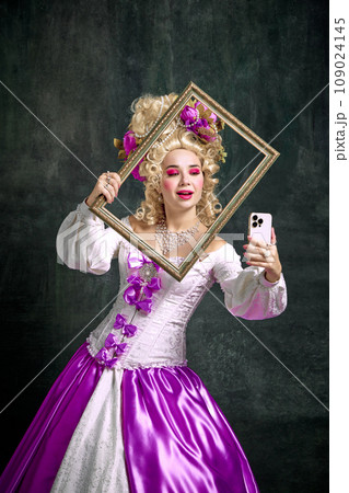 Woman dressed like classic historical character, in old-fashioned dress holding retro frame and taking selfie on mobile phone against vintage background. Woman dressed like classic historical character, in old-fashioned dress holding retro frame and taking selfie on mobile phone against vintage background. 109024145