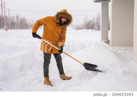 A man cleans and clears the snow in front of the house on frosty day. Cleaning the street from snow on a winter day. Snowfall and severe snowstorm in winter. 109025095