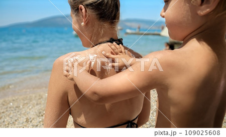 Cute baby boy lovingly applies sunscreen lotion to his mother's back on a sunny sea beach. Skin protection and caring for loved ones Cute baby boy lovingly applies sunscreen lotion to his mother's back on a sunny sea beach. Skin protection and caring for loved ones 109025608