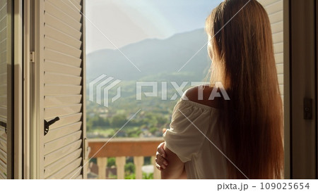 Young brunette woman opens window blinds at her villa and walking out on the terrace or balcony. Concept of summertime, enjoying mountain view, holiday and travel 109025654