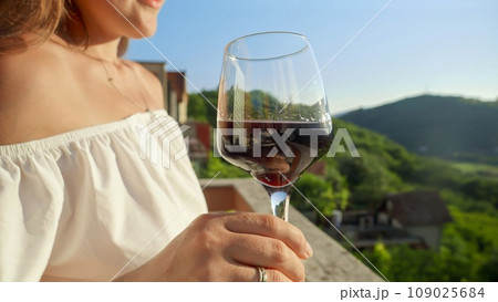 Young woman with glass of red wine gracefully held, merges with the magic of a mesmerizing sunset and mountain view from the villa balcony or terrace. Travel, summertime, and holiday serenity 109025684