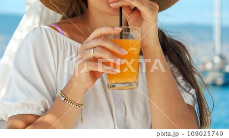 Closeup of elegant woman holding glass of orange juice and drinking it through straw while relaxing on wooden sea pier 109025749
