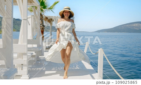 Young Woman in a Flowing Light Dress and Straw Hat Joyfully Runs along a Wooden Pier at the Beach, Embodying the Essence of a Relaxing Summertime Vacation Young Woman in a Flowing Light Dress and Straw Hat Joyfully Runs along a Wooden Pier at the Beach, Embodying the Essence of a Relaxing Summertime Vacation 109025865