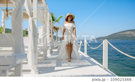 Smiling young woman, in a light dress and chic straw hat, walks on rustic wooden pier by the sea, infusing fashion with the allure of coastal bliss Smiling young woman, in a light dress and chic straw hat, walks on rustic wooden pier by the sea, infusing fashion with the allure of coastal bliss 109025866