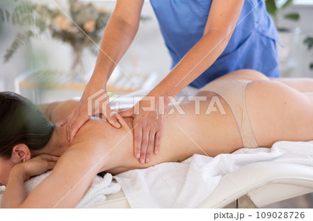 Relaxed young woman lying on massage table enjoying remedial body massage done by professional masseur in modern spa salon 109028726