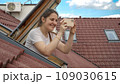 Young smiling woman looking out of the open attic window and making photos on her smartphone. Concept of tourism, holiday in Europe, travel and journey 109030615