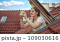 Young woman, with a bright smile, uses her smartphone to capture photos while looking out of an open attic window. Tourism, holidaying in Europe, and the spirit of travel and exploration 109030616